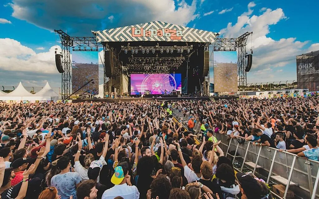 Foto: plateia de mão levantada com palco no fundo e céu azul