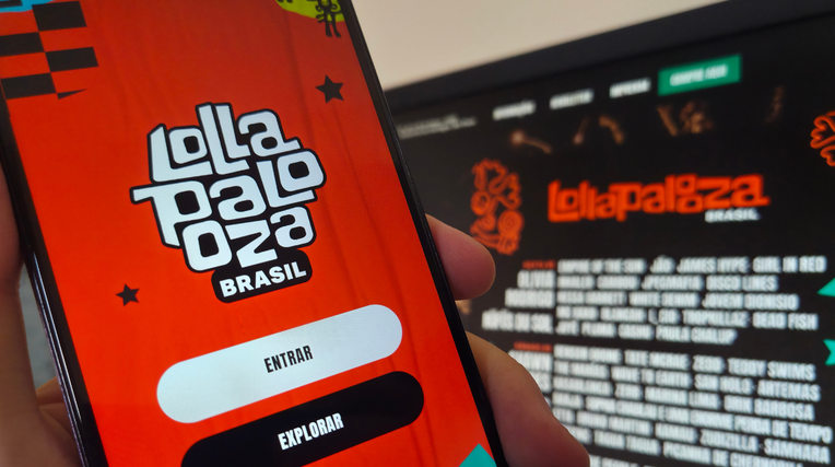 Foto: uma mão segurando um celular com a logo do evento lollapalooza na tela, ao fundo, desfocado, a grade de cantores que estarão no evento