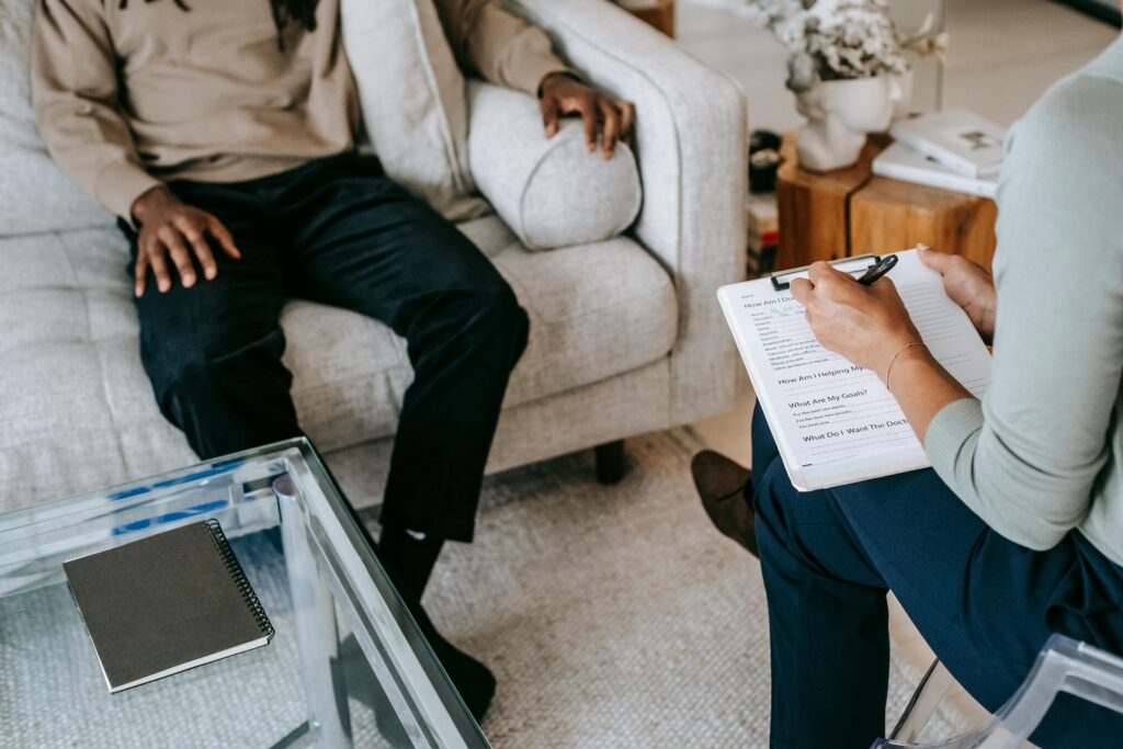 Um homem sentado em um sofá, com uma psicóloga na sua frente, segurando um caderno, fazendo anotações da sessão de terapia.