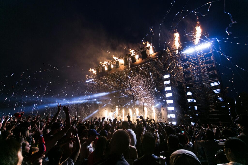 Foto: palco de show ao fundo com muitas luzes e plateia mais em frente com braços levantados.