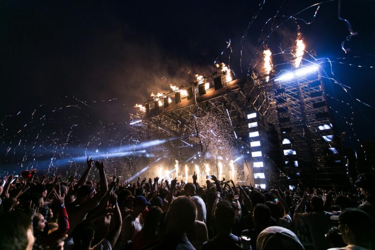 Foto: palco de show ao fundo com muitas luzes e plateia mais em frente com braços levantados.