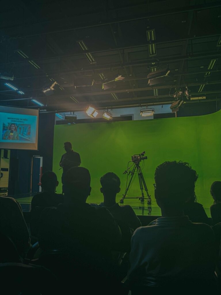 Um grupo de pessoas assiste a uma apresentação em um estúdio equipado com chroma key verde. Em primeiro plano, o público está em silhueta, enquanto ao fundo um palestrante segura um microfone próximo a uma câmera profissional. Há também um telão com a imagem de uma repórter e a legenda “Microfone de Mão (Direcional)”. O ambiente está escuro, com foco na iluminação de estúdio e no conteúdo técnico apresentado.