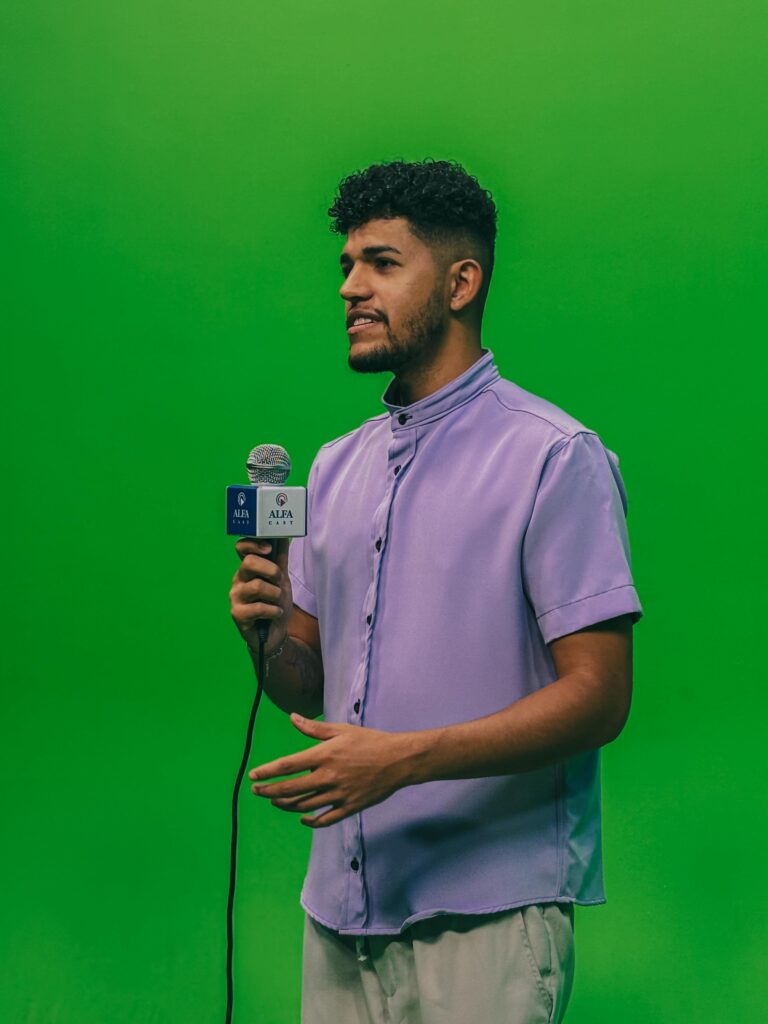 Estudante de camisa lilás segura o microfone da Alfa e se comunica com expressão confiante. Está posicionado em frente ao fundo verde do estúdio, reforçando o cenário típico de gravações televisivas.