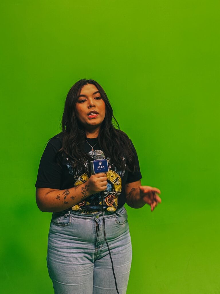 Estudante segura um microfone com a logo da Alfa e fala diante de um fundo verde de chroma key. Ela gesticula com uma das mãos enquanto se expressa, demonstrando concentração e domínio do espaço de gravação.