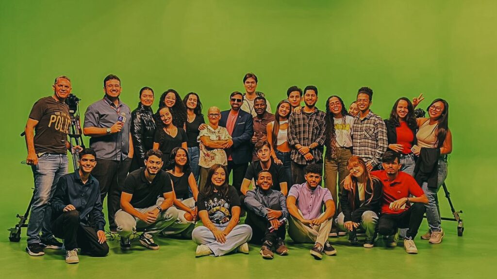 Turma de estudantes de Jornalismo da Unialfa, unidade Bueno, posa para foto coletiva em estúdio de TV com fundo verde (chroma key), na unidade Perimetral. Ao todo, cerca de 30 pessoas, entre alunos e professores, estão reunidas sorrindo, com parte do grupo sentado no chão e os demais em pé. Alguns seguram equipamentos como câmeras e microfones, representando a prática jornalística vivenciada durante a aula.