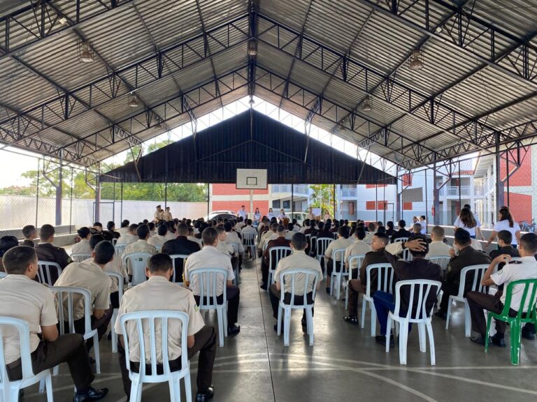 Imagem de alunos sentados em cadeiras de plástico na cor branca, em uma quadra de futebol. Ao fundo, o Time Acolher, grupo que realizou a ação.